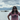 Woman wearing a pink Rock Life t-shirt and sunglasses, standing with hands on hips in front of a scenic Kodiak coastline with rocky islands, blue water, and mountains under a cloudy sky — island-inspired women's adventure apparel