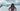 Woman wearing a pink Rock Life t-shirt and sunglasses, standing with hands on hips in front of a scenic Kodiak coastline with rocky islands, blue water, and mountains under a cloudy sky — island-inspired women's adventure apparel