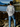Person standing on a rocky shore facing the water, wearing a light gray Rock Life long sleeve tee with a scenic design on the back. Mountains and trees are visible in the background.