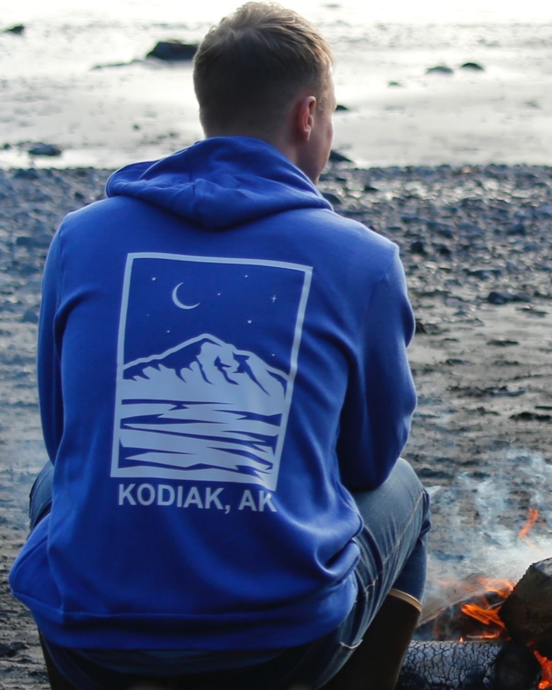 Person wearing blue Rock Life mountain hoodie sitting by campfire on Kodiak beach in Alaska