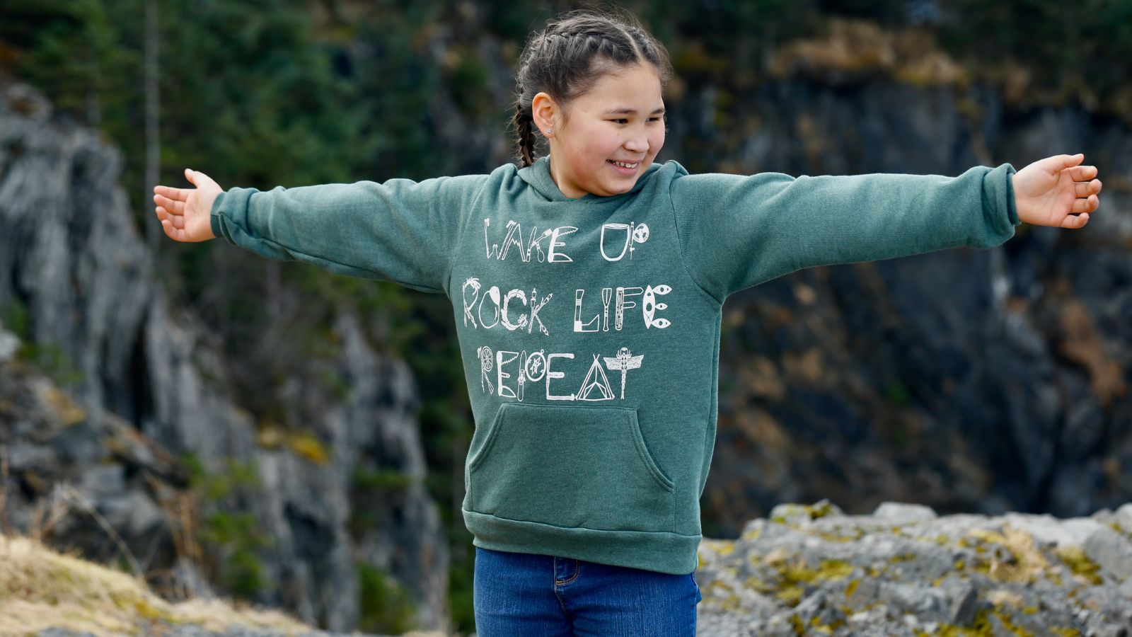 Child wearing green Adventure in Every Letter youth hoodie with arms outstretched in Kodiak, Alaska