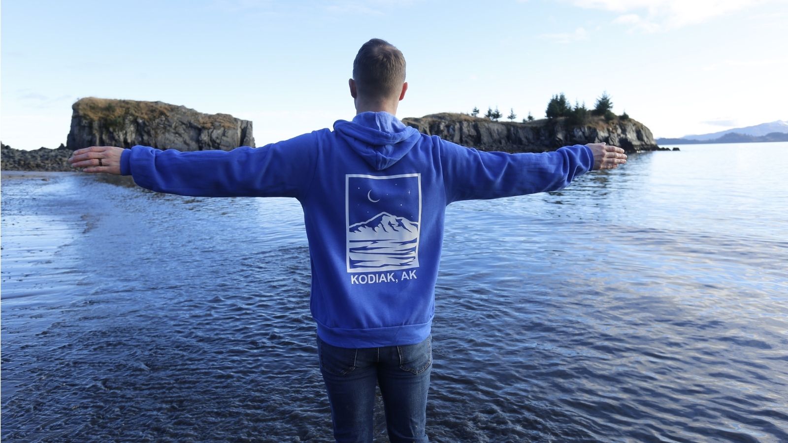 Person wearing blue hoodie with mountain graphic and Kodiak, AK text standing by water with scenic Alaska background